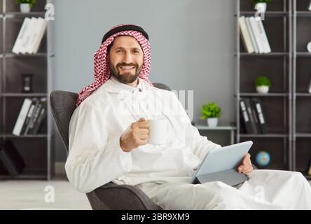 Arabischer Mann, der während der Fernarbeit eine Kaffeepause macht, posiert mit einer Tasse Kaffee, einem digitalen Tablet Stockfoto