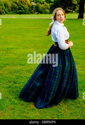 Eine Frau in weißem Hemd, braunem Gürtel und langem kariertem Rock steht auf einem grasbewachsenen Feld und blickt über ihre Schulter. Stockfoto