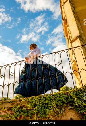 Eine Frau in traditionellem schottischem Outfit steht auf einem Balkon und blickt in den Himmel. Sie hält ein Buch und lehnt sich auf das Geländer. Stockfoto