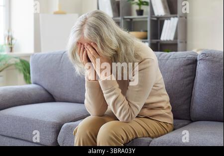 Alte Seniorin sitzt drinnen auf dem Sofa in Not zu Hause, bedeckt das Gesicht mit den Händen und weint Stockfoto