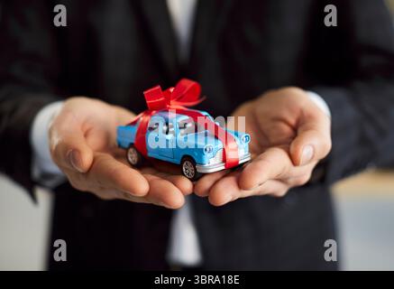 Nahaufnahme eines Verkäufers in formellem schwarzem Anzug, der ein kleines Spielzeugauto mit roter Geschenkschleife in den Händen hält. Stockfoto