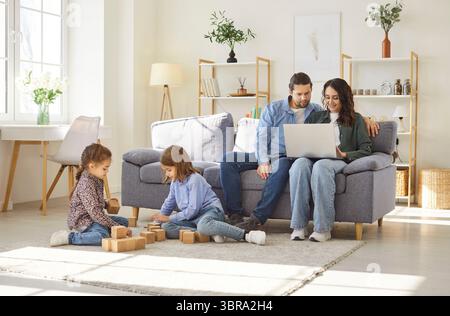 Familie zu Hause, Tochter und Sohn spielen auf dem Boden mit Würfeln, Eltern sitzen auf dem Sofa und legen sich am pc aus Stockfoto