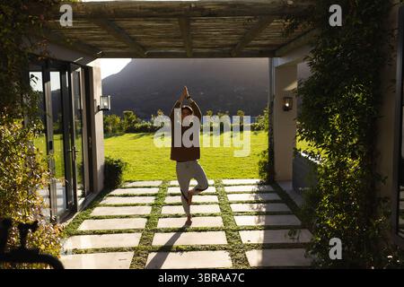 Asiatischer Mann, der Yoga auf der Terrasse unter hölzerner Pergola neben einer Glasschiebetür praktiziert Stockfoto