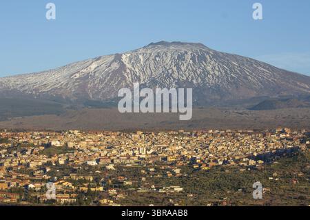 Blick aus der Vogelperspektive auf sonnendurchflutete Gebäude, eingebettet vor den majestätischen, schneebedeckten Hängen des Ätna, wo Schatten über die Landschaft tanzen, Catania, Ital Stockfoto