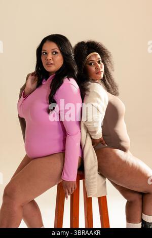 Verschiedene weibliche Models mit Strickjacke und Stirnband, die im Studio Rücken an Rücken auf Holzhocker sitzen Stockfoto