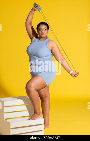 Asiatische Frau in blauer Sportbekleidung mit Fuß auf Kiste und Sprungseil in gelbem Studio Stockfoto