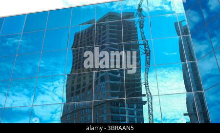 Hellblaues Glas spiegelt die hoch aufragende Baustelle und Wolken darüber wider. Stockfoto