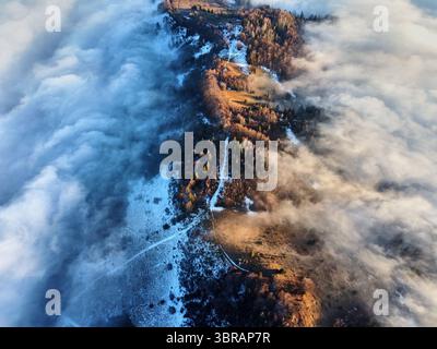 Aus der Vogelperspektive der nebelbedeckten Landschaft, in der das Sonnenlicht durch Wolken eindringt und eine Mischung aus Land und dichtem Wald offenbart. Schatten von Bäumen und Nebel erzeugen ein dramatisches Zusammenspiel von Licht und Dunkelheit. Stockfoto