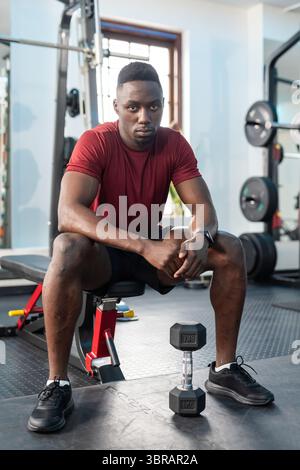 Afroamerikaner, der auf einer verstellbaren Bank im Fitnessstudio sitzt und ein kastanienbraunes Hemd trägt, der eine Hexenhantel hält Stockfoto