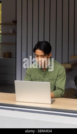 Asiatischer Mann mit olivgrünem Hemd, der am Laptop sitzt und im Büro an der Holztheke sitzt Stockfoto