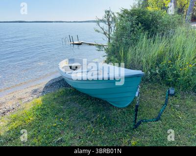 Ein kleines blaublaues Boot ruht an einem grasbewachsenen Ufer mit Blick auf einen ruhigen See an einem sonnigen Tag. Stockfoto