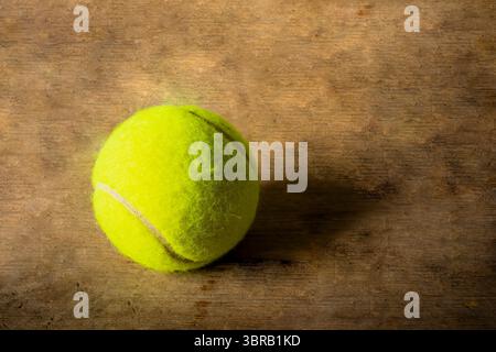 Ein hellgelber Tennisball hebt sich auf einer schön strukturierten rustikalen Holzoberfläche ab, die von einem warmen, gerichteten Licht beleuchtet wird. Stockfoto