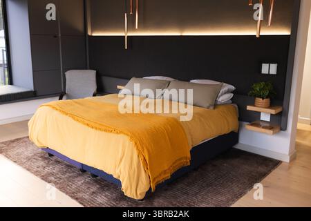 Kingsize-Bett mit Senfdecke, grauen Kissen und Kupferanhängern an Holzkohlewand im Schlafzimmer Stockfoto