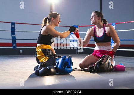 Zwei weibliche Boxer kniend und mit Fäusten zwischen Handschuhen, Kopfbedeckung, Wasserflasche im Boxring Stockfoto