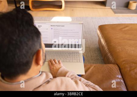 Asiatischer Mann, der auf einem braunen Sofa im Wohnzimmer sitzt und auf silbernem Laptop schreibt, Kopierraum Stockfoto