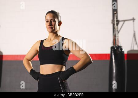 Sportlerin in Sportbekleidung stehend mit Händen auf Hüften neben hängendem Boxsack im Fitnessstudio Stockfoto