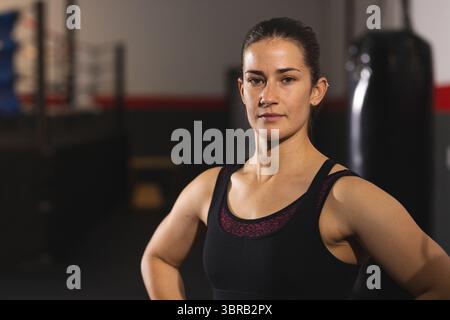 Frau mit schwarzem Sport-Top-Arm akimbo im Dojo mit Boxring und Boxsack Stockfoto