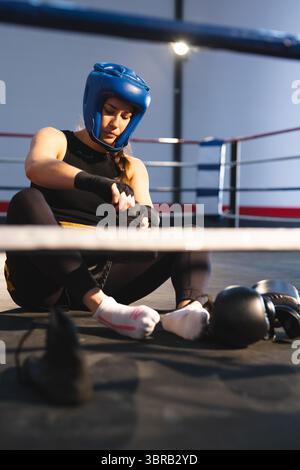 Weibliche Boxerin sitzt im Boxring und trägt einen blauen Helm, der Hände mit schwarzen Wickeln in der Nähe von Handschuhen umwickelt Stockfoto
