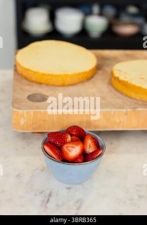Keramikschale mit halbierten Erdbeeren auf der Arbeitsplatte neben dem Schneidebrett mit Biskuitschichten Stockfoto