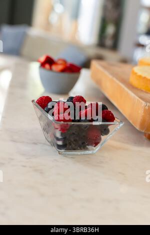 Quadratische Glasschale mit Himbeeren und Heidelbeeren auf Marmorarbeitsfläche in der Nähe einer grauen Keramikschale Stockfoto