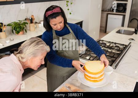Verschiedene weibliche Freunde stapeln Schwammkuchenschichten auf weißem Teller an der Marmorküche Stockfoto