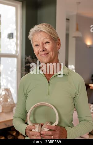 Ältere Frau mit beigefarbenen Kopfhörern im Wohnbereich mit Korblampe und getrockneten Blumen Stockfoto