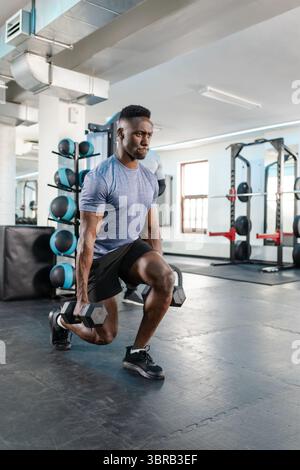 Ein afroamerikanischer Mann, der gewichtete Ausfallschritte mit Hexen-Kurzhanteln auf Gummiboden im Fitnessstudio durchführt Stockfoto