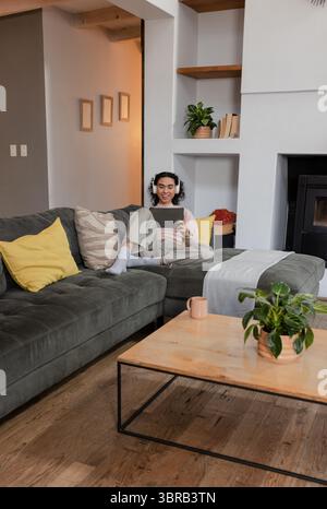Nicht binäre Person mit schnurlosen Kopfhörern, die mit einem Tablet auf einem Ecksofa im Wohnzimmer sitzt Stockfoto