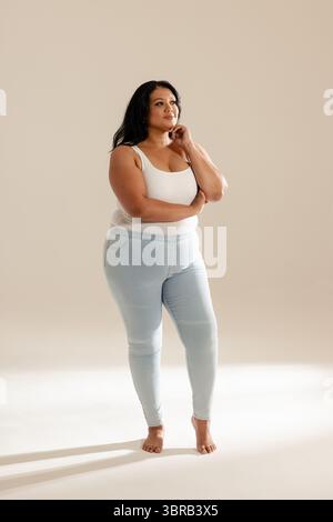 Frau, die Arme überkreuzte Hand auf dem Kinn in einem Studio in neutralem Ton trägt, in weißem Oberteil und Jeans Stockfoto