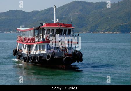 Eine kleine schwarz-weiß-rote Fähre zwischen den Inseln in Hongkong. Das Schiff ist mit den Hügeln von Lantau im Hintergrund zu sehen. Stockfoto
