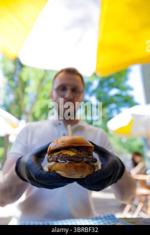 Köstlicher, handgefertigter Burger, der vom Koch präsentiert wird, mit frischen Zutaten und leckeren Toppings unter einem hellen Outdoor Canopy in einem lebhaften Dining Stockfoto