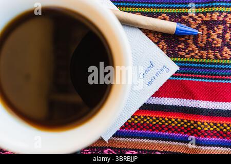 Kaffeetasse mit einer Nachricht, die einen guten Morgen auf einem farbenfrohen Tisch mit Inka-Gewebe sagt. Stockfoto
