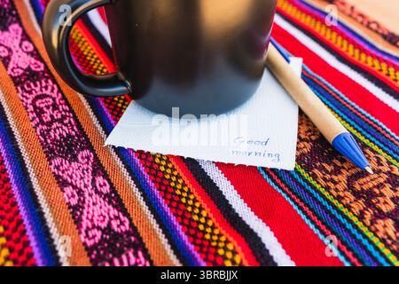 Kaffeetasse mit einer Nachricht, die einen guten Morgen auf einem farbenfrohen Tisch mit Inka-Gewebe sagt. Stockfoto