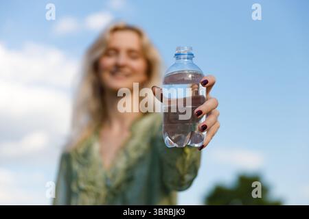 Eine junge Frau hält freudig eine klare Plastikflasche Wasser vor einem wunderschönen blauen Himmel mit weichen Wolken, die Erfrischung und Entspannung betont Stockfoto