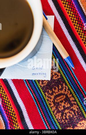 Kaffeetasse mit einer Nachricht, die einen guten Morgen auf einem farbenfrohen Tisch mit Inka-Gewebe sagt. Stockfoto