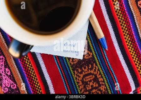 Kaffeetasse mit einer Nachricht, die einen guten Morgen auf einem farbenfrohen Tisch mit Inka-Gewebe sagt. Stockfoto