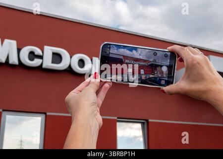 McDonalds-Fassade wird mit Smartphone fotografiert 10.07.25, Limburg an der Lahn: Symbolfoto, Illustrationsbild, Symbolbild, Illustrationsfoto, Alltagsszene zwei Hände halten ein Smartphone und fotografieren die Fassade eines McDonald s-Restaurants mit dem markanten weißen Logo. Der Fokus liegt auf dem Display des Smartphones, das Szene wiedergibt. Die rote Wand des Gebäudes und Teile eines Werbebanners mit Eis und Pommes sind im Hintergrund sichtbar. Die Aufnahme vermittelt ein modernes, urbanes Konsumerlebnis und die allgegenwärtige Smartphone-Nutzung. Eisenbach Hessen Deutschland *** Mc Stockfoto