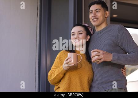 Verschiedene Paare stehen auf dem Balkon in der Nähe der Glasschiebetür und halten Keramikbecher, während sie nach rechts schauen Stockfoto