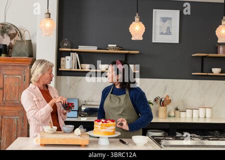 Verschiedene ältere Freundinnen bereiten auf der Kücheninsel mit Mischschalen einen mehrschichtigen Obstkuchen zu Stockfoto
