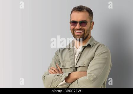 Porträt eines selbstbewussten lächelnden Mannes, der eine Sonnenbrille mit überkreuzten Armen trägt und vor einem neutralen Hintergrund steht. Lässiger Stil und positiver Ausdruck. Stockfoto