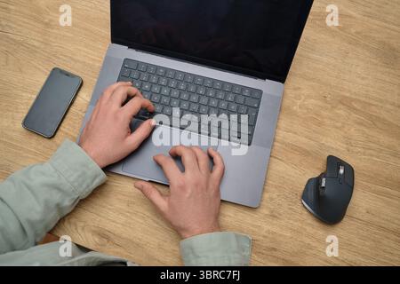 Nahaufnahme einer Person, die auf einer Laptop-Tastatur an einem Holzschreibtisch tippt, mit Smartphone und ergonomischer Maus in der Nähe. Einrichtung des produktiven Arbeitsbereichs. Stockfoto