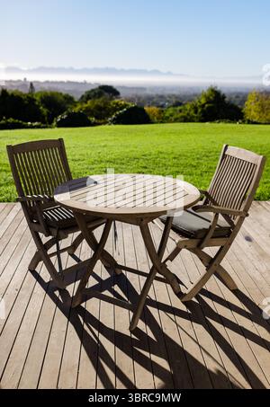 Runder Holztisch und Stühle auf Holzterrasse mit Blick auf grünen Rasen und nebelige Berge Stockfoto