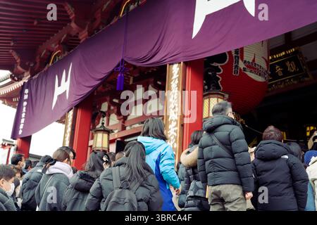 Besucher steigen die Stufen in den Senso Ji Tempel unter tiefvioletten Bannern hinauf Stockfoto