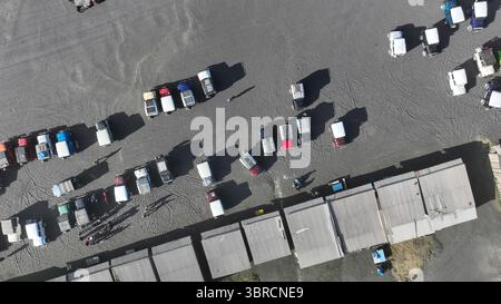 Von oben parken Reihen klassischer Jeeps neben rustikalen Marktständen, die lange Schatten auf den grauen vulkanischen Sand werfen. Stockfoto