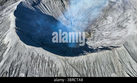 Ein faszinierender Blick aus der Luft in das Herz eines vulkanischen Kraters, wo sich der blaue Rauch tief in der rauen grauen Landschaft erhebt. Natur RAW Stockfoto