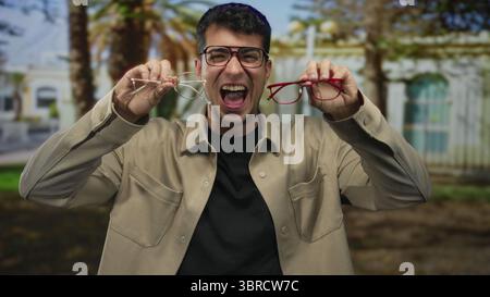 Ein Mann, der eine Brille trägt, während er zwei Paare im Park vergleicht, zeigt, wie junge hispanische Männer Brillen bewerten, um die Wahl des Rahmens zu treffen. Stockfoto