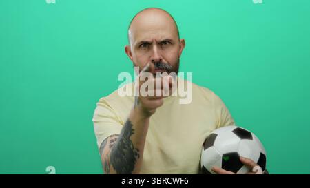 Bärtiger kahlköpfiger Mann mit Tätowierungen, der Fußball hält, bedrohliche Gesten vor isoliertem grünen Hintergrund macht, Emotionen und Intensität ausdrückt Stockfoto