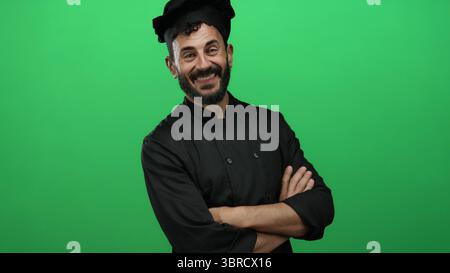 Lächelnder hispanischer männlicher Koch mit Bart in schwarzer Uniform, der selbstbewusst vor lebendigem grünem Hintergrund steht und kulinarisches Know-how zeigt. Stockfoto