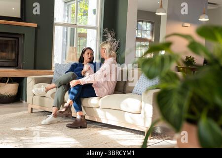 Verschiedene Freundinnen sitzen auf dem Sofa im Wohnzimmer und unterhalten sich, während sie Keramikbecher halten Stockfoto