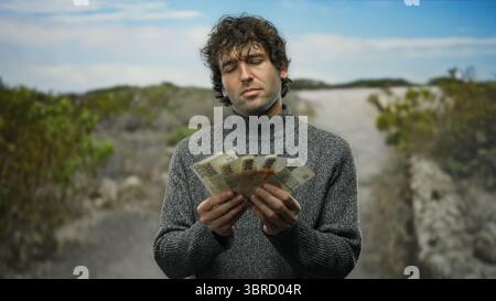 Hispanischer Mann, der tschechische Kronen-Banknoten im Freien in einem Park zählt, umgeben von einer natürlichen Landschaft und trägt einen gemütlichen Pullover, der die finanziellen Banknoten hervorhebt Stockfoto